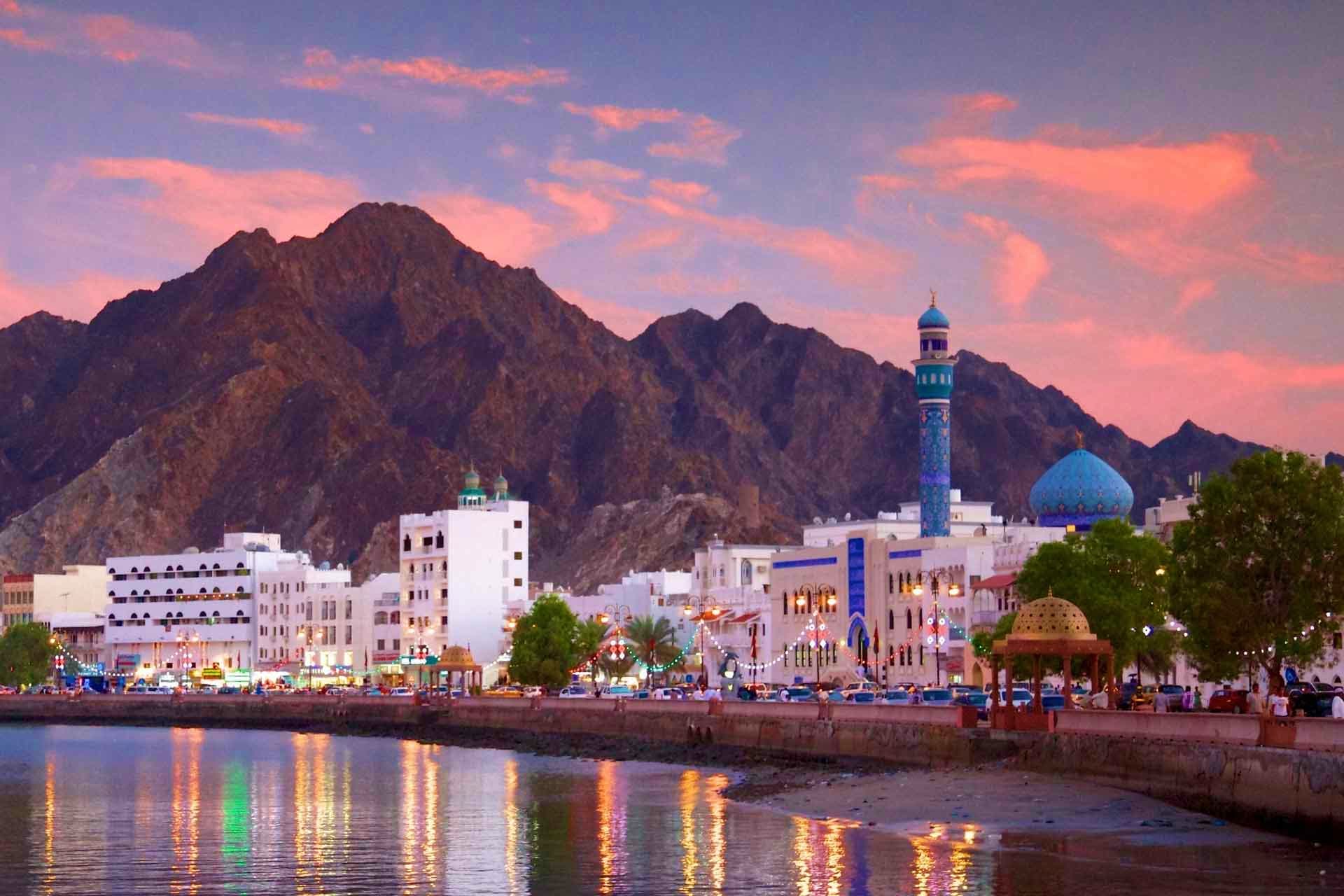 Oman Visa Muscat waterfront at sunset with white buildings, blue-domed mosque, minaret, and rocky mountains reflected in calm water